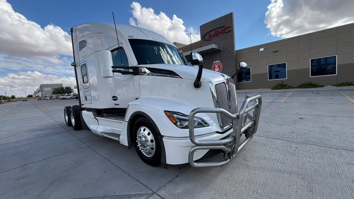 Truck front-left view of a white 2024 Kenworth T680 with chrome bull bar and grille guard in a dealership lot.