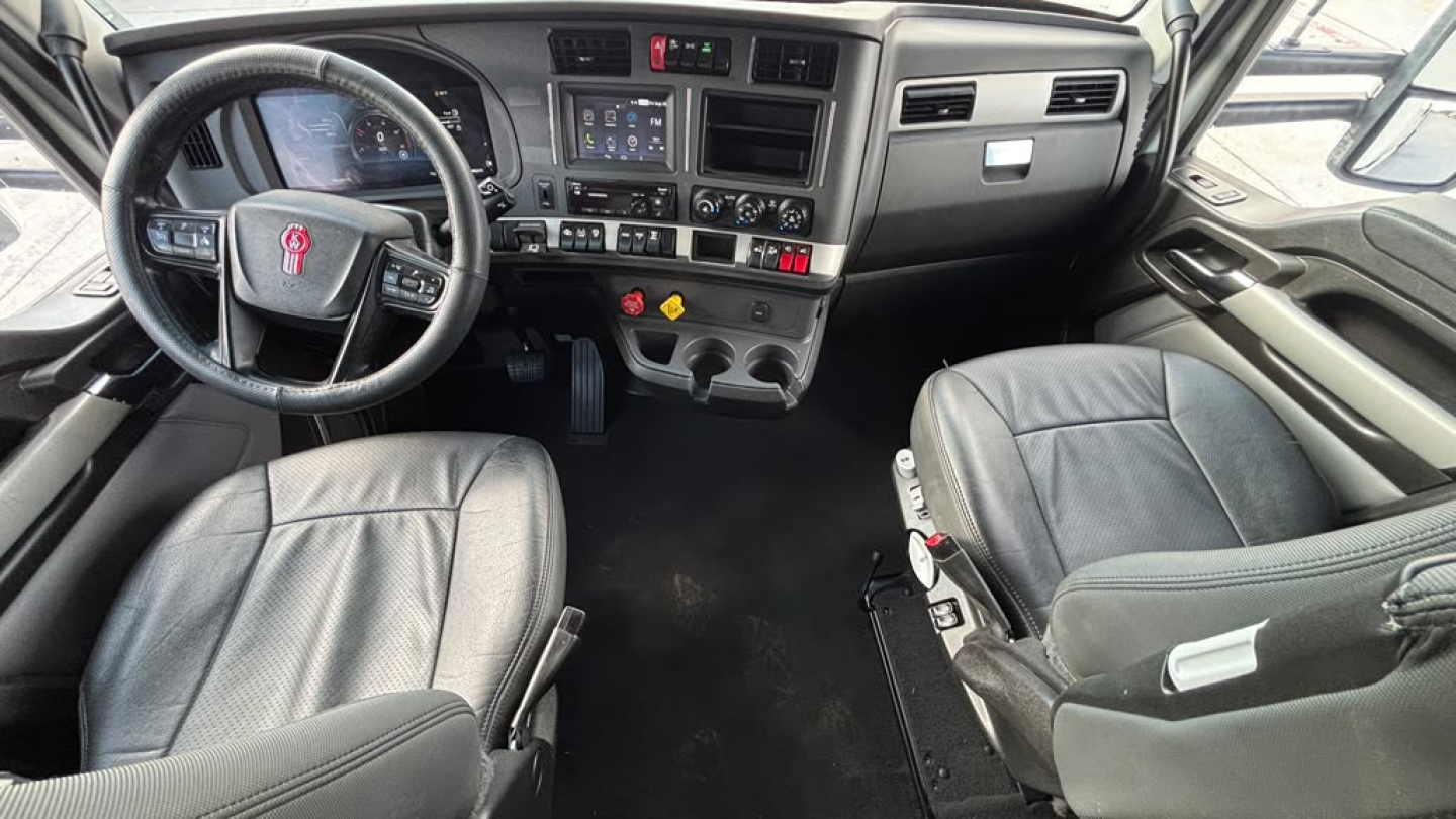 Cab interior of a 2024 Kenworth T680 showing steering wheel, dashboard, center console with digital screen, and two seats in view.