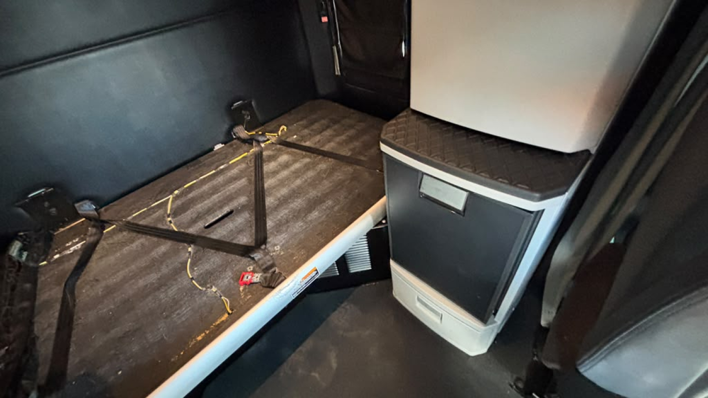 Kenworth T680 sleeper cab interior showing side countertop and storage fridge area with seat belt hardware and metallic floor.