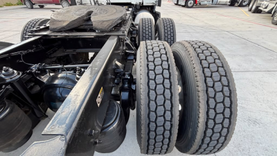 Truck chassis with dual rear axles shown in close-up view; visible components include frame rails, air tanks, drivetrain, and suspension. No cabin, engine details visible; tires show tread pattern.