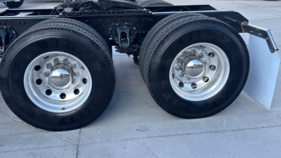 Kenworth T680 drivetrain dual rear wheels close-up showing polished aluminum wheels and tires, suspension components, chassis, and mud flap mounting.