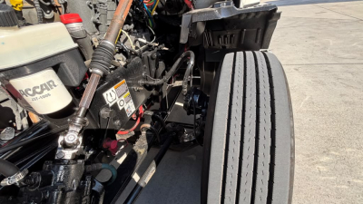 Truck engine bay area with various hoses and components; visible labels include a white reservoir with 'OCAR' text and numbers, and a black battery/electrical area with warning labels.
