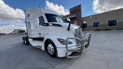 Truck front-left view of a white 2024 Kenworth T680 with chrome bull bar and grille guard in a dealership lot.
