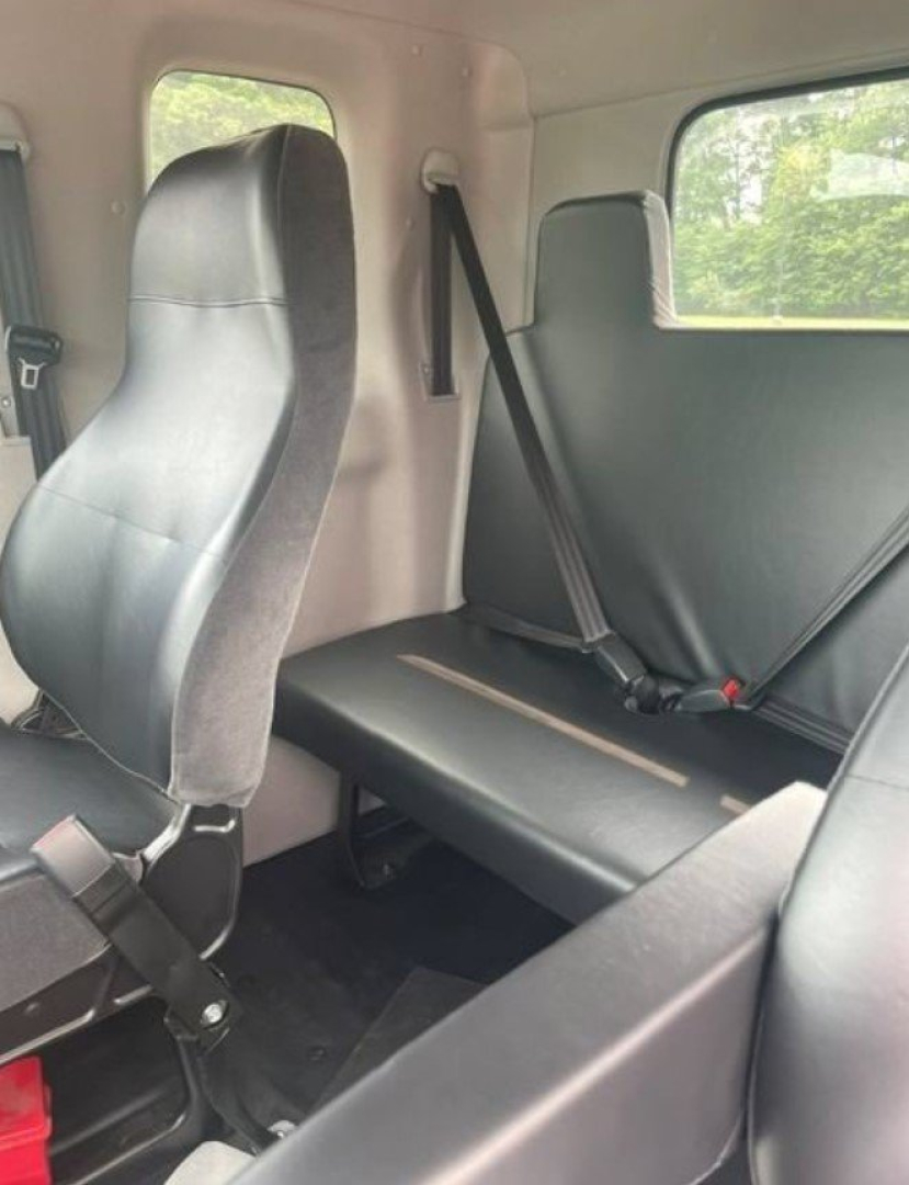 Cab interior showing two-person seating with seat belts and a bench seat in the rear, black upholstery, side window, and light-colored panels.