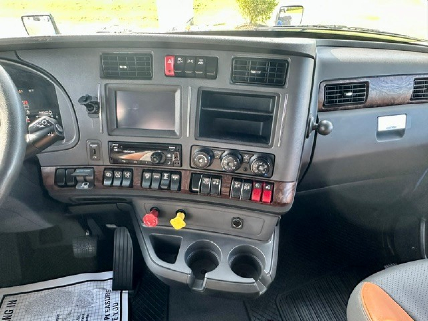 Kennworth T680 truck dashboard view showing infotainment screens, control switches, cup holders, and yellow/red hazard knobs.