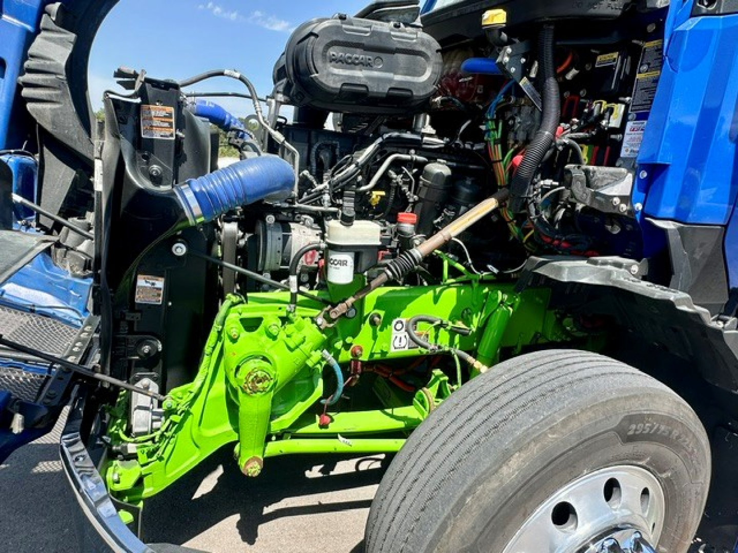 Kenworth T680 engine bay showing green front axle and blue cab with various hoses and components visible.
