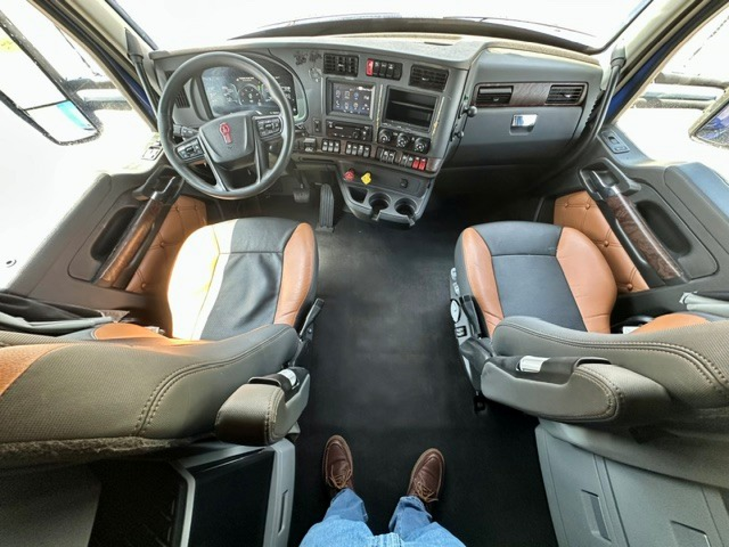 Interior of a Kenworth T680 cab showing steering wheel, dashboard with controls, two-tone brown and black seats, and captain chairs with leg room.