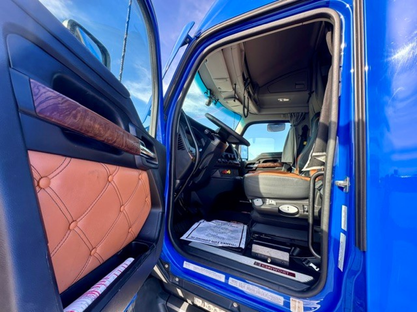 Truck cab interior visible through open door: blue exterior, tan leather-like door panel, steering wheel, dashboard, driver's seat, overhead controls; shows interior view of Kenworth truck cabin.