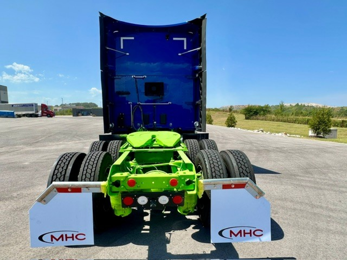 Kenworth truck rear view showing bright green fifth wheel assembly and white mudflaps with MHC logo; blue cab back and clear sky.