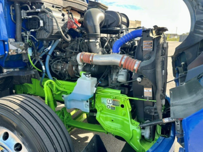 Kenworth T680 engine bay showing coolant reservoir, blue hoses, green-painted frame rail, and various hoses and components.