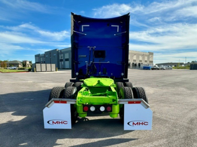 Kenworth T680 rear view showing bright green chassis and white MHC mud flaps, blue dump-style truck bed raised, dual rear axles, clear sky.