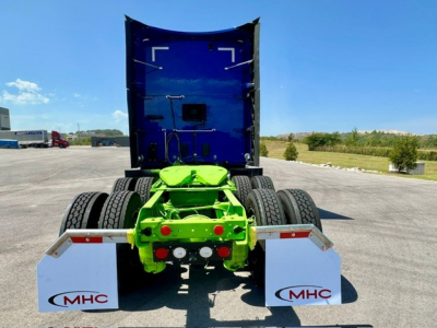 Kenworth T680 rear view showing green chassis, dual rear axles, white mud flaps with MHC logo, and blue sleeper cab in background.