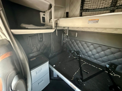 Truck sleeper interior with quilted black side panels, small storage cabinet with two drawers, overhead bunk area, and cargo straps showing in a Kenworth T680 cabin.