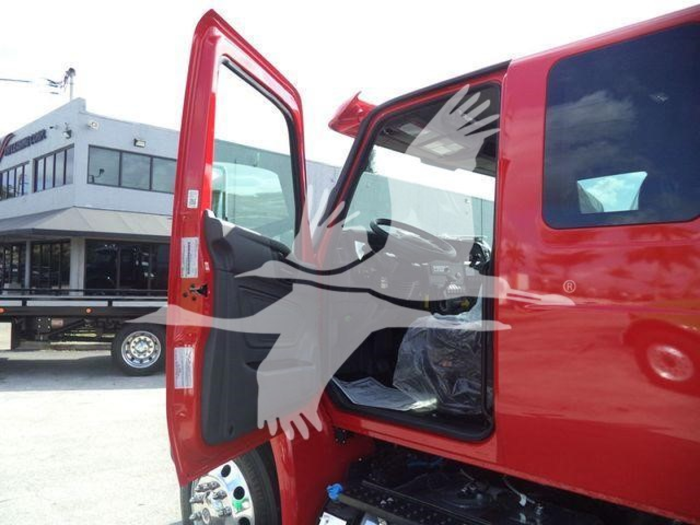 Truck cab with open passenger door showing interior and side window; red exterior, black step; background shows dealership lot and building.