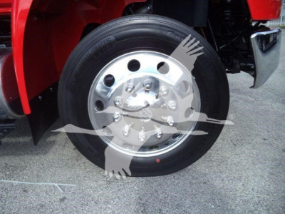 Wheel and hub of a heavy-duty truck front axle showing a polished aluminum hub with multiple lug bolts and black tire, red fender in background.