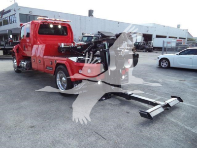 Tow truck with a mounted winch in a lot, red International MV truck visible, crane/winch equipment and black arms, industrial building in background.