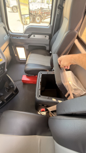 Truck cab interior with open center console storage box and seat in Mack PI64T. Visible doors, cabin seating, and storage compartment lid partially lifted.