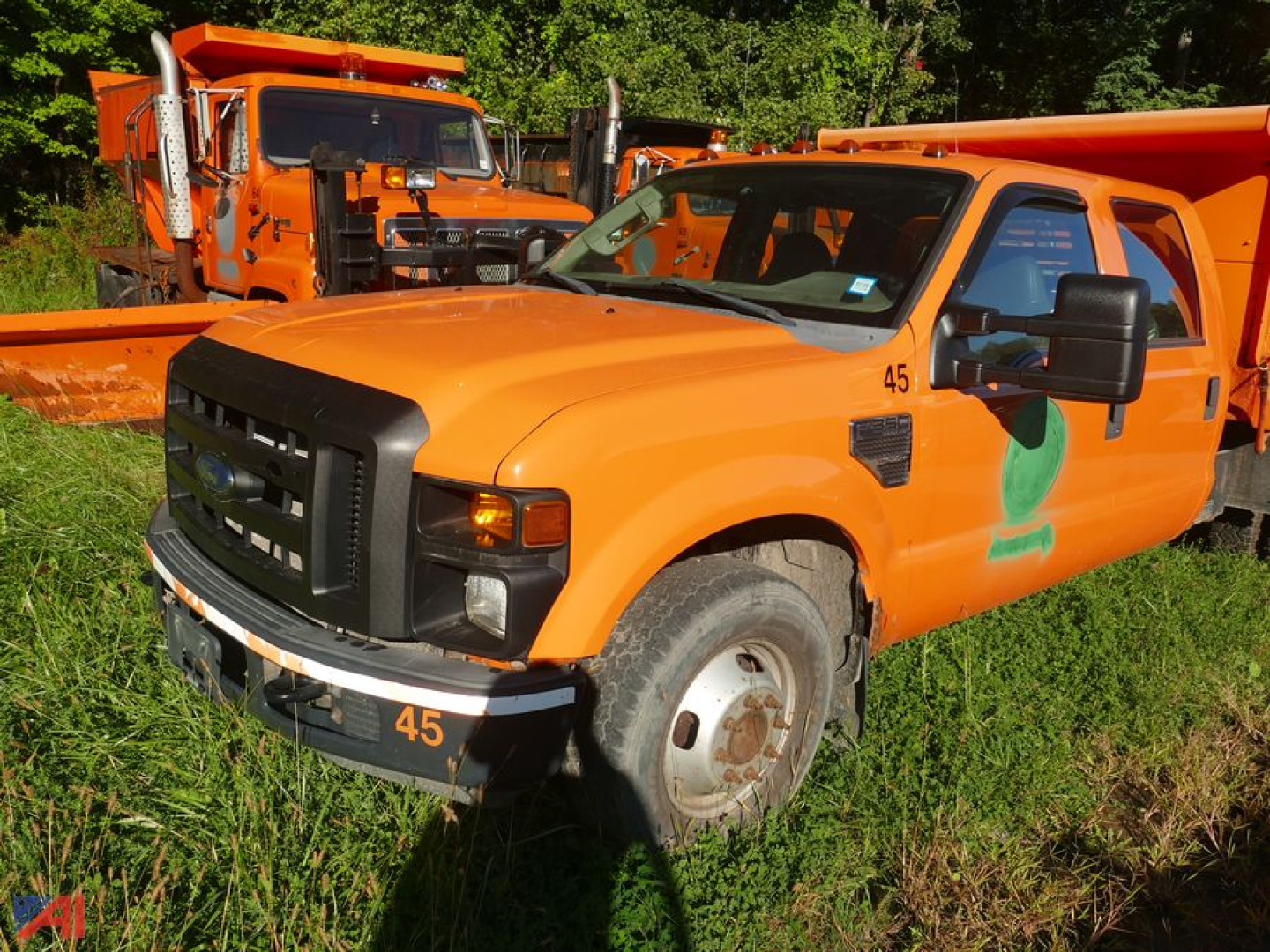 Ford F-350 Super Duty cab and chassis with orange paint, dual rear wheel, front grille, headlights, and side mirrors; vehicle in grassy lot with another orange truck in background.