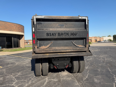 Rear view of a dump truck bed on a 2023 Peterbilt 567 showing the tailgate with 'STAY BACK 300'' text and dual rear axles, dirt on metal surface, parking lot background