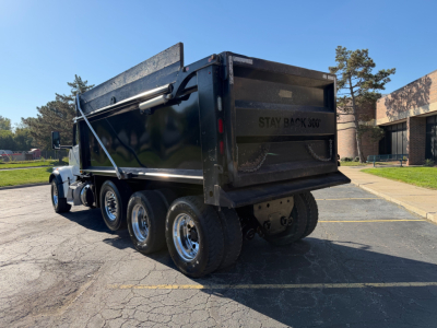 Dump truck rear and side view showing Peterbilt 567 dump bed with black paint and dual rear axles; visible tires and chrome rims; tailgate area with 'STAY BACK' text.