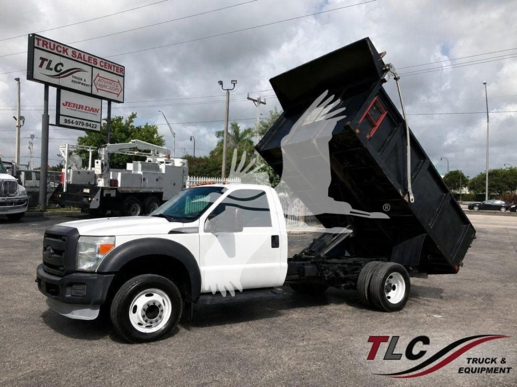Dump truck in a tilt-bed configuration with white cab and black dumping bed raised, Ford emblem on grille, TLC Truck & Equipment signage in background.
