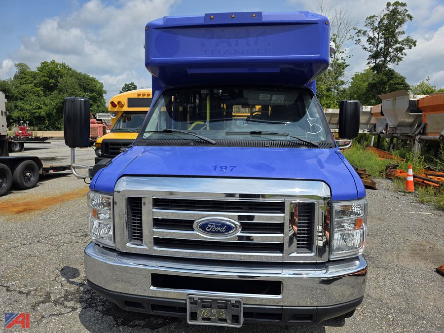Front view of a blue 2018 Ford E-Series passenger bus with a raised blue hood, chrome grille, and Ford emblem; multiple orange school buses in background.