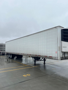 Refrigerated trailer by a loading dock, white Wabash reefer with a front refrigeration unit, side panels, and dual rear axles on a wet pavement.