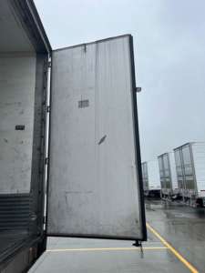 Rear of refrigerated trailer door open showing interior and outside parking area with other trailers in background.