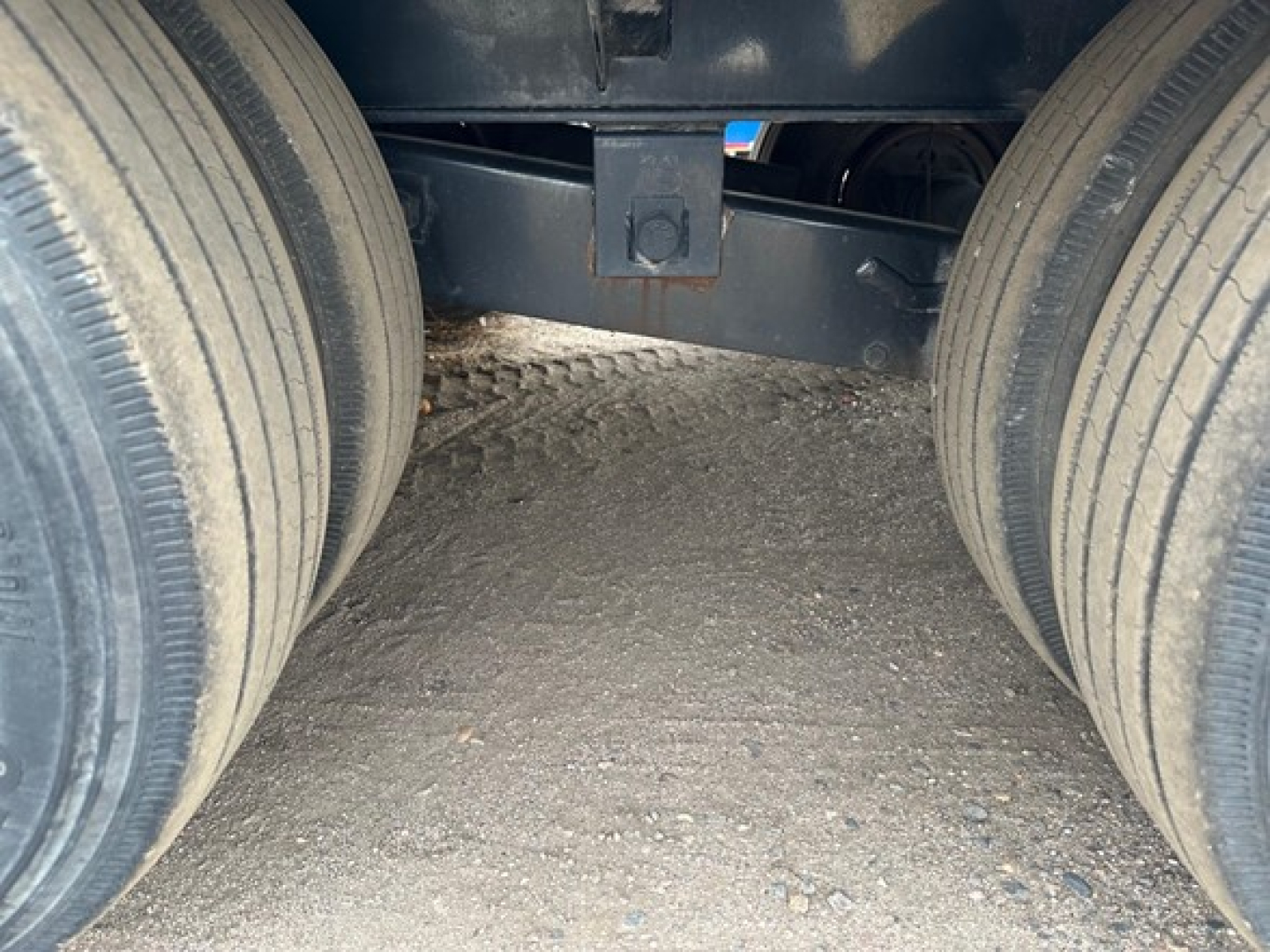 Trailer axle assembly view showing dual rear tires and undercarriage of a flatbed trailer; dark chassis and ground surface visible.