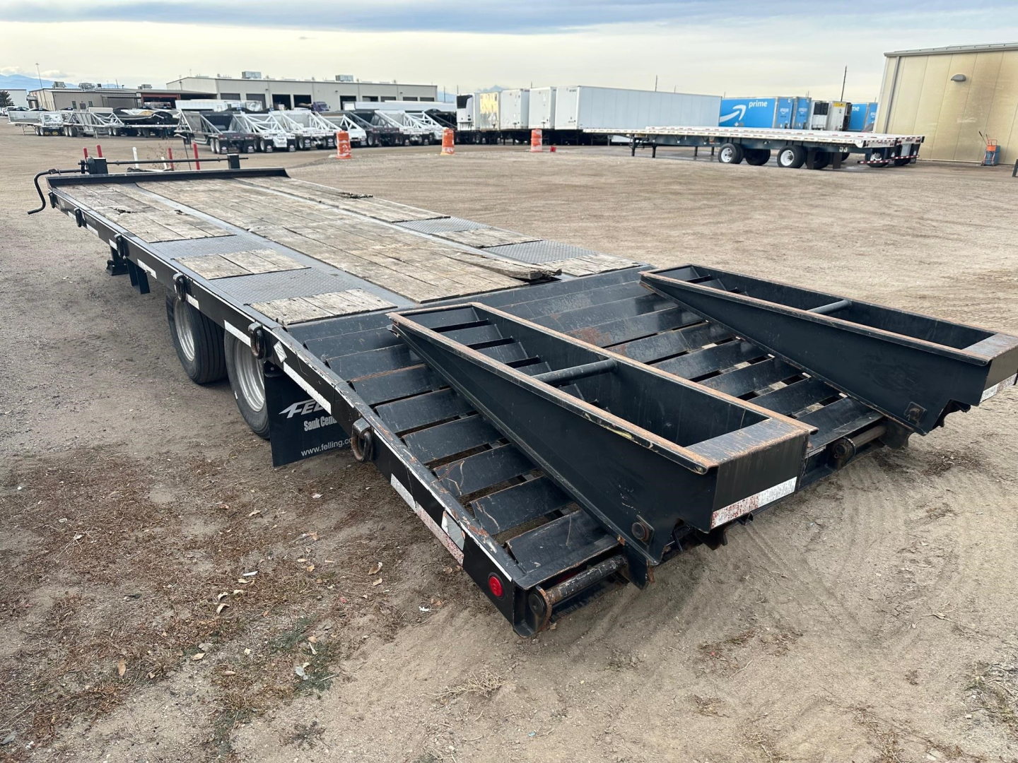 Flatbed trailer with Felling branding visible, side rails and loading ramps folded, showing wear on decking and tires. Background shows industrial lot with trucks and buildings.