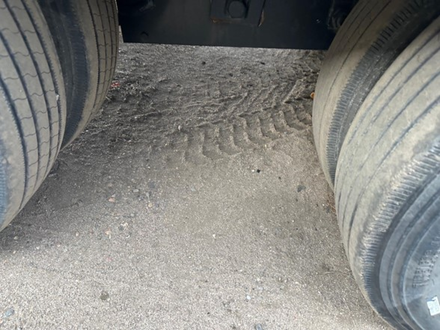 Back-end view of a flatbed trailer hitch area between dual rear tires, showing gravel and tire treads.