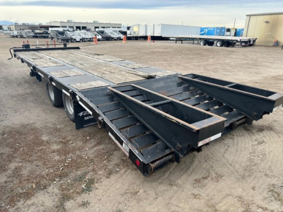 Flatbed trailer with long black steel frame and side ramps, dual rear axles, visible wear and dirt on deck, parked in a gravel lot with other trailers in background.
