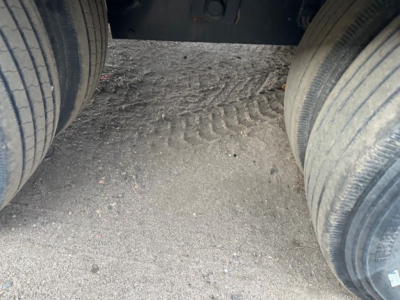 Back-end view of a flatbed trailer hitch area between dual rear tires, showing gravel and tire treads.