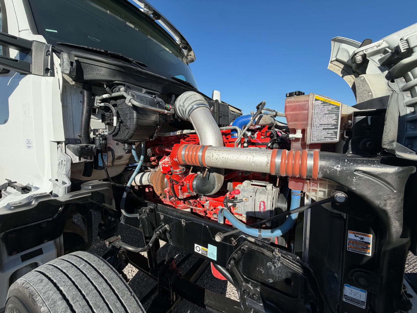Kenworth T880 engine bay showing red engine block and connected hoses; view of front wheel well and radiator area; blue coolant lines and exhaust components visible.