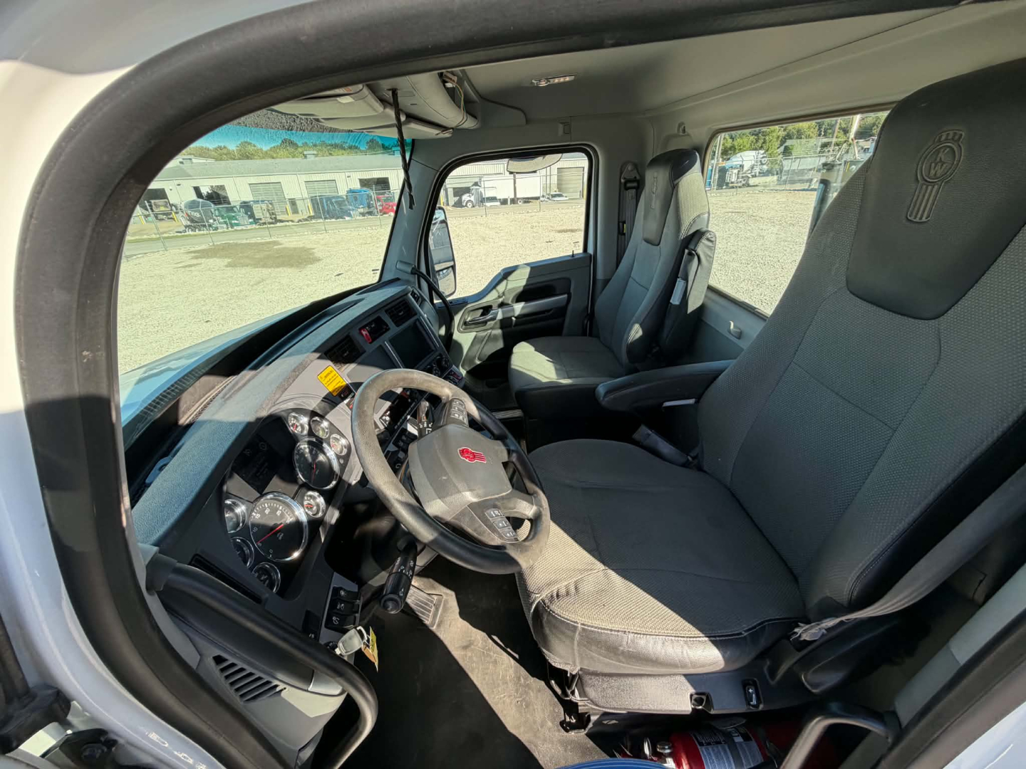 Kenworth T880 cab interior showing steering wheel and driver seat with gauges visible; dashboard and windshield; gray fabric seats.