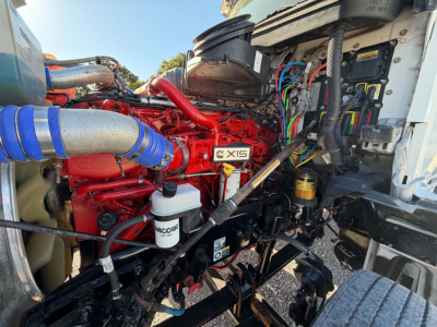 Kenworth T880 engine bay with red Cummins X15 engine, blue intake hose, various hoses and wiring, and white coolant reservoir visible.