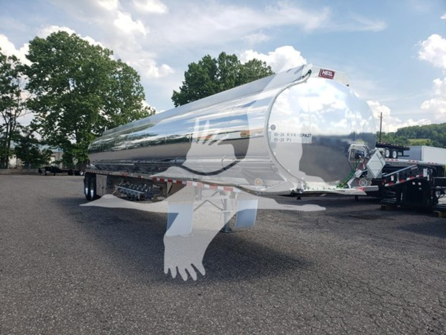 Trailer: polished aluminum Heil 406A Petro tanker trailer in yard, rear door and dual axles visible, reflection on body, set against trees and cloudy sky.