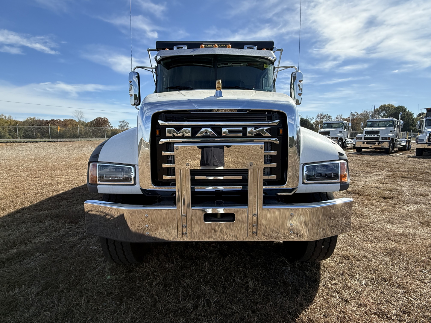 Front view of a white Mack dump truck with large chrome bumper and grille, 'MACK' lettering on the grille. Visible headlamps and side mirrors. Parking lot background with other trucks.