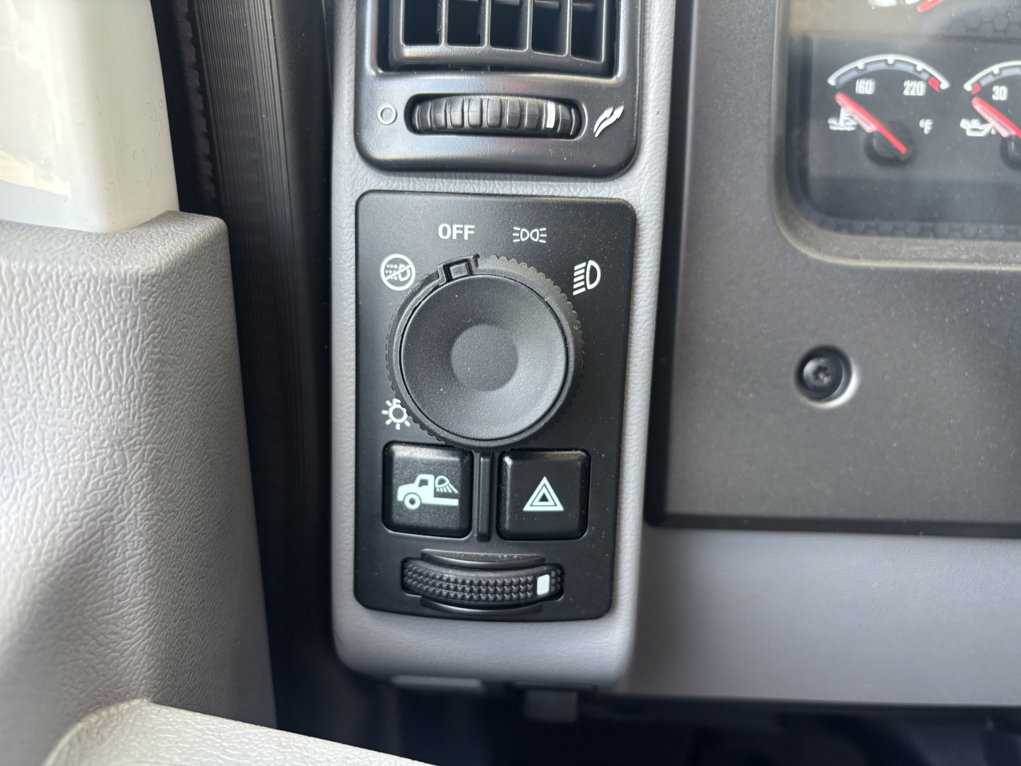 Dashboard controls and headlight switch area in a new Mack GR64B. The image shows a circular dial with OFF and headlight icons, small push buttons below for tow/auxiliary functions, and a portion of the instrument cluster on the right.