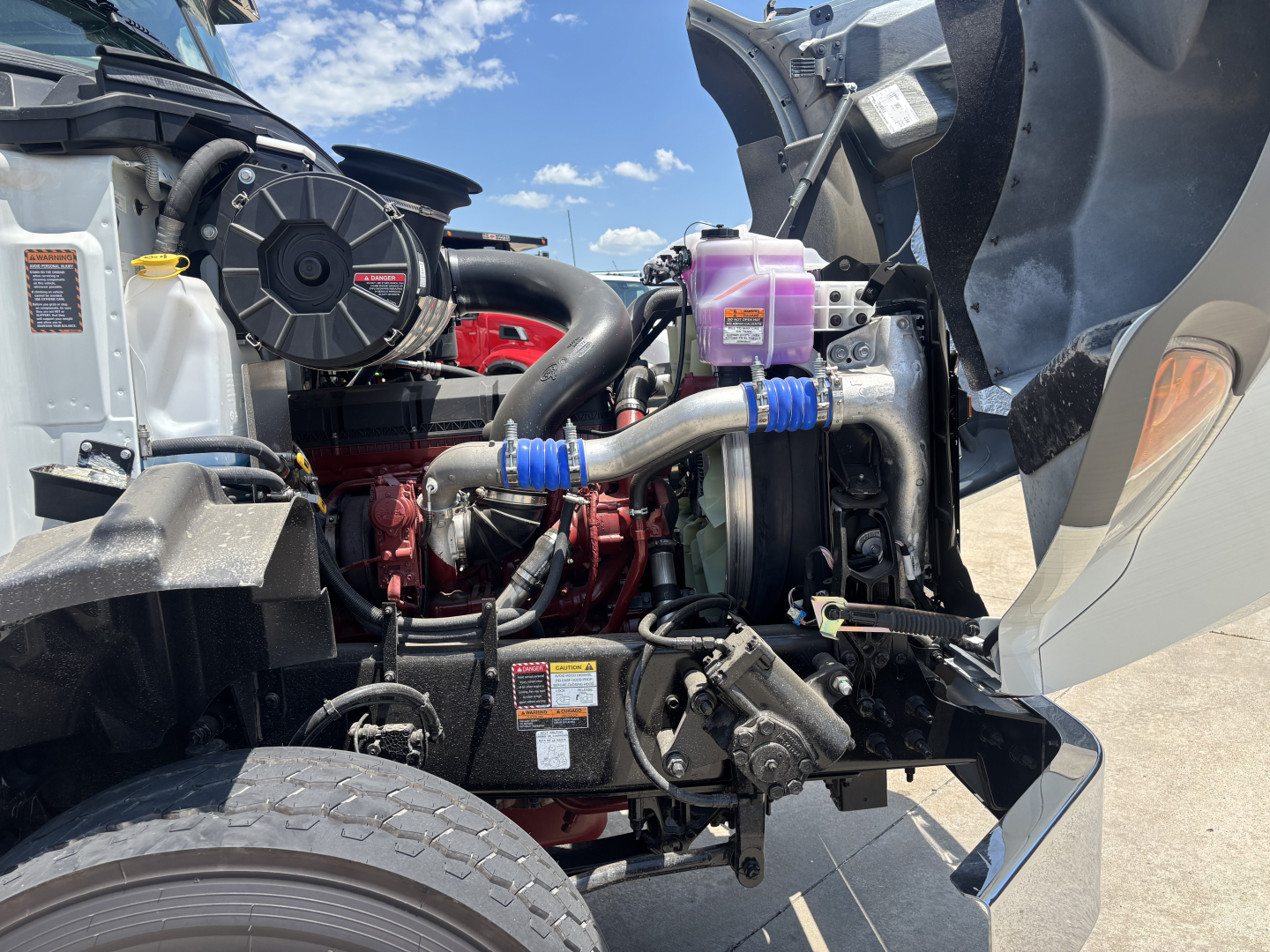 Mack GR64B engine bay with cooling system, hoses, and purple coolant reservoir cap visible; front wheel and components in view.