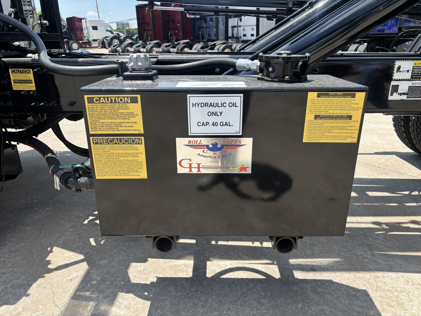 Black rectangular hydraulic oil reservoir with yellow caution and precaution labels on sides, capped at top, with two discharge pipes at bottom and a central white label reading HYDRAULIC OIL ONLY CAP. 40 GAL. and Roll Offs logo.