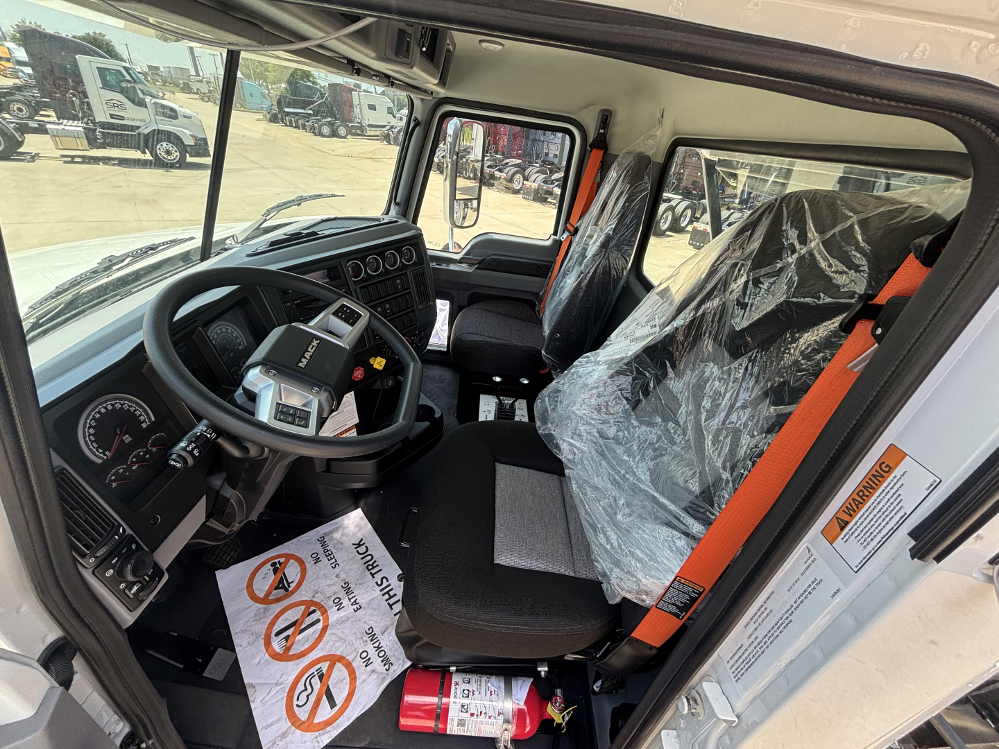 Interior of a 2026 Mack GR64B roll-off truck cab showing steering wheel, dashboard, and seat with plastic cover; visible warning labels on the door frame and orange seatbelt.