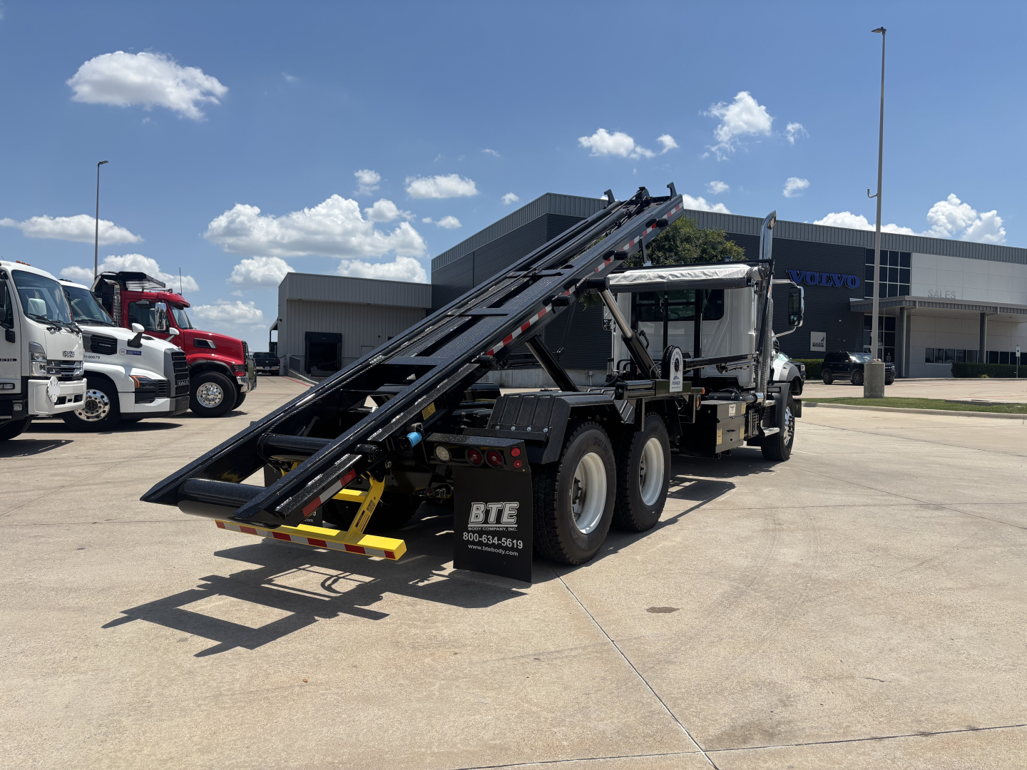 Mack GR64B roll-off truck in service bay lot showing rear loading ramp and chain system.