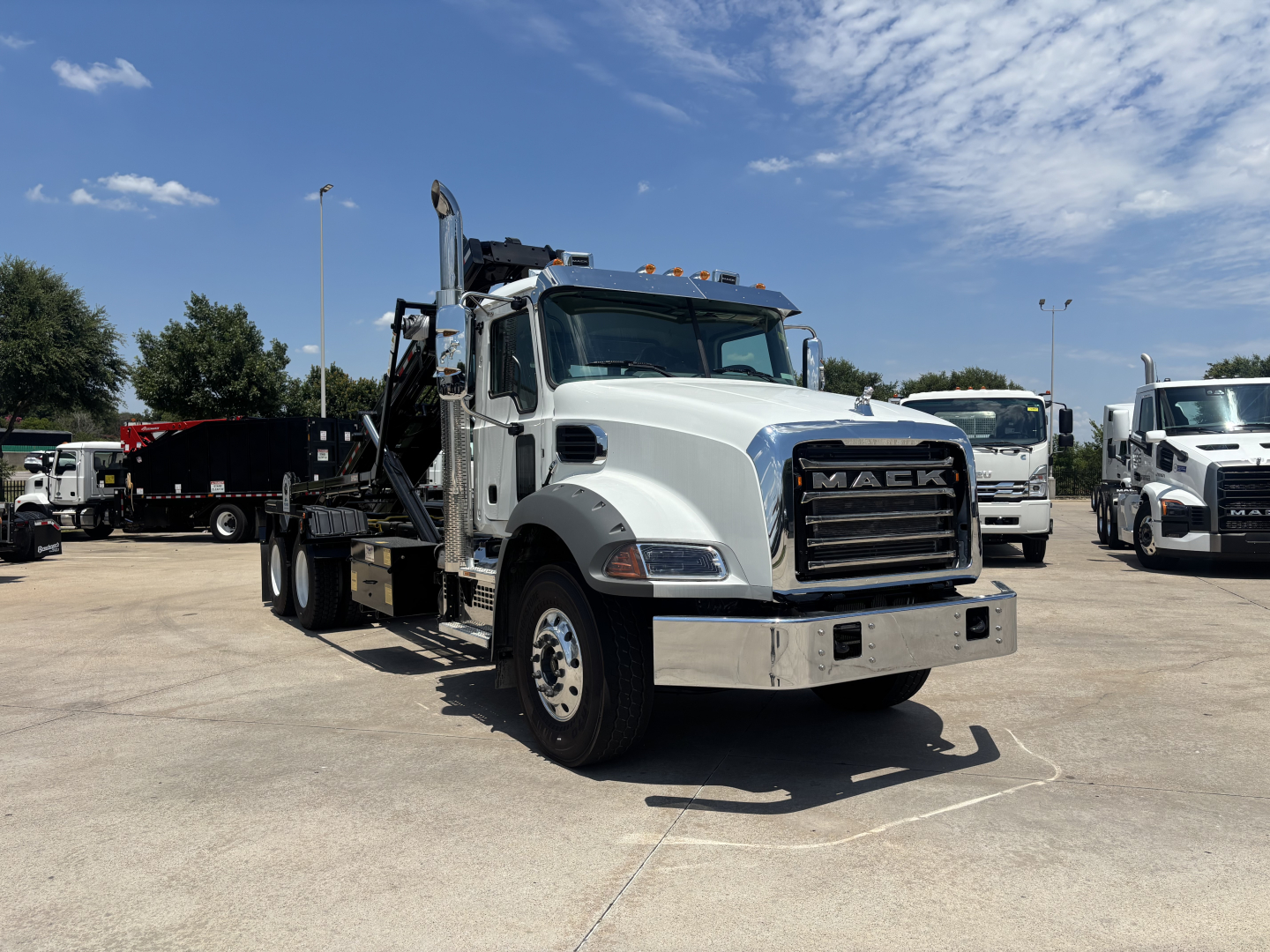 Front-left view of a white 2026 Mack GR64B rolloff truck in a dealership lot, showing the crane and flatbed with chrome bumper and large grille.