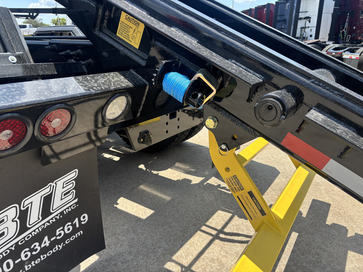 Truck bed area showing hydraulic hook-up and blue protective sleeve on fuel line near yellow support leg and warning label in foreground.