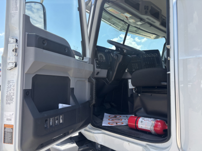 Truck cab interior of a 2026 Mack GR64B roll-off truck showing the driver seat, dashboard, open door with storage pockets, and a fire extinguisher on the floor.