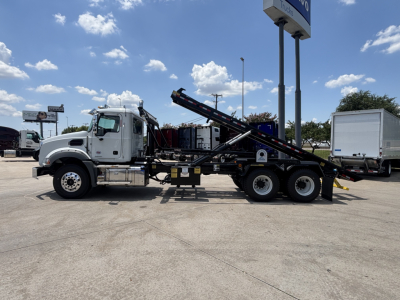 2026 Mack GR64B rolloff truck side view showing hydraulic systems and tilting bed, white cab, black chassis, in-service label details not legible.