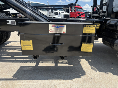 Rolloff truck rear showing a black toolbox with yellow caution labels and a central handle; sticker with blue and red logo and text; surrounding trucks in background.