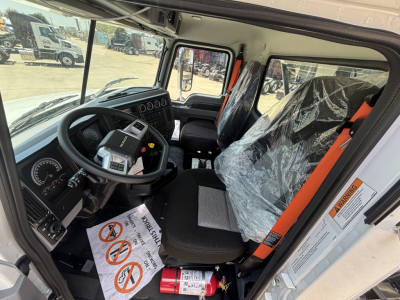 Interior of a 2026 Mack GR64B roll-off truck cab showing steering wheel, dashboard, and seat with plastic cover; visible warning labels on the door frame and orange seatbelt.