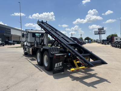 Mack GR64B rolloff truck in a dealership lot, side view showing hydraulic ramp/loader bed and stacked dual rear axles.